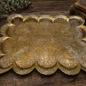 May include: A rectangular brass tray with a scalloped edge and an intricate, etched floral pattern across the surface. The metal has a weathered, aged patina with visible texture and varying shades of gold and dark brown throughout the design.
