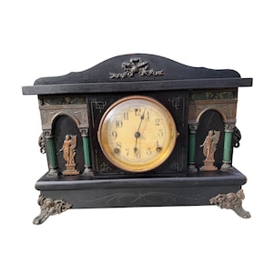 May include: A black mantel clock with a central circular dial featuring black Arabic numerals and hands. The clock case is decorated with green columns, two bronze-toned relief figures, and a bow motif at the top, supported by ornate feet.