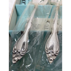 May include: Two silver-toned metal spoons with ornate, embossed scrollwork patterns on the handles. The spoons are encased in clear plastic protective sleeves and are positioned inside a light blue cardboard box with visible text.