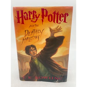 Puede incluir: Un libro de tapa dura titulado Harry Potter and the Deathly Hallows de J.K. Rowling. La portada presenta una ilustración de una persona de cabello oscuro con una túnica oscura y una camisa verde, extendiendo la mano hacia un cielo naranja y amarillo.