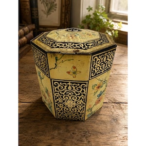May include: An octagonal metal storage tin with a cream-colored base featuring painted floral branches, green leaves, and orange butterflies. The tin has alternating panels decorated with intricate black and white scrollwork patterns and a matching lid.