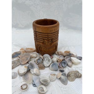May include: A brown wooden mug carved with a stylized tiki face featuring horizontal lines and a wide, toothy grin. The mug is surrounded by a collection of various small, natural seashells in shades of white, gray, tan, and brown.