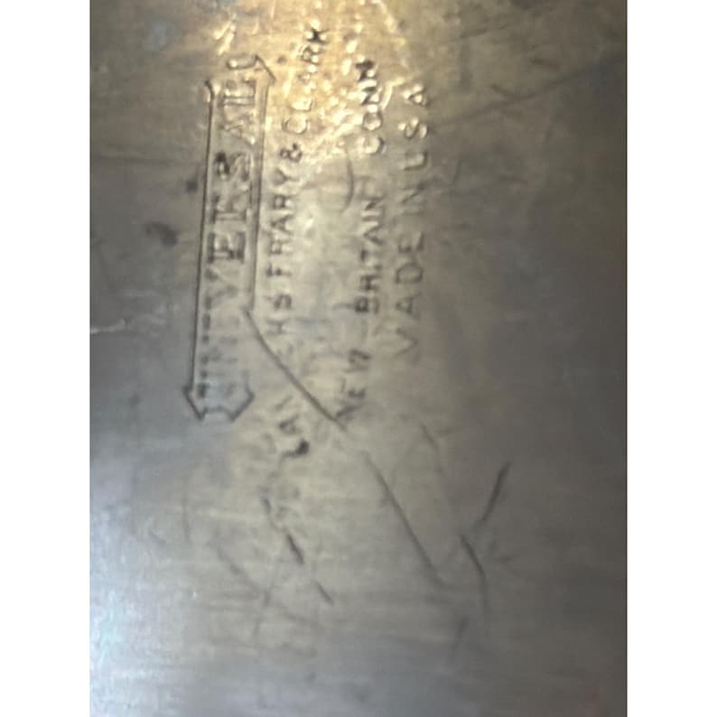May include: A metallic surface featuring an embossed stamp that reads UNIVERSAL, LANDERS FRARY & CLARK, NEW BRITAIN CONN, MADE IN USA. The surface shows visible scratches and signs of wear across the textured, light-coloured metal material.
