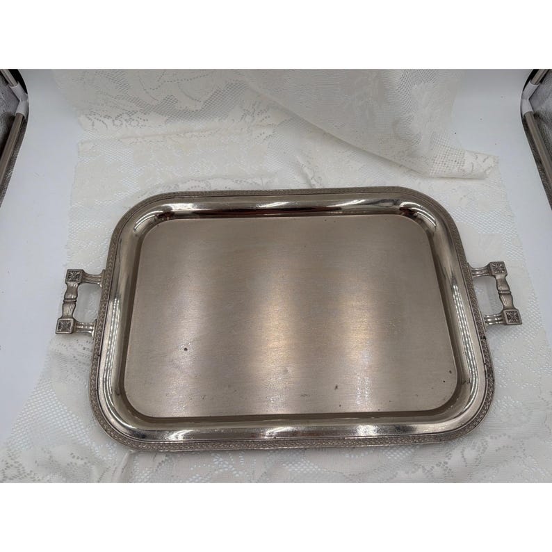 May include: A rectangular silver-toned serving tray with rounded corners and a polished, reflective surface. The tray features a decorative border with a textured pattern and two ornate, rectangular handles on opposite sides.