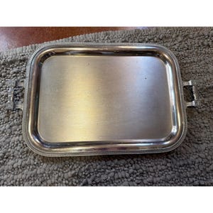 May include: A rectangular silver-toned serving tray with a smooth, reflective central surface and a raised border featuring a decorative rope-like pattern. The tray includes two metallic handles on opposite sides with a segmented design.