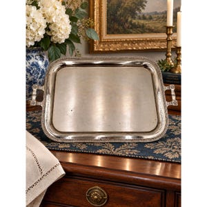 May include: A rectangular silver-toned serving tray featuring a polished metallic surface, a raised beaded border, and two ornate handles on the sides. The tray has a smooth, reflective finish and a structured frame with decorative detailing on the handles.