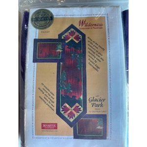 May include: A fabric kit for a table runner and placemats, featuring a wilderness design. The kit includes fabric for the runner top, binding, and two matching placemats. The design incorporates geometric shapes and images of trees.