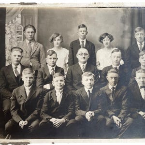May include: A vintage black and white photograph of a group of people, likely a class or club. The subjects are dressed in formal attire, including suits, ties, and dresses. The image has a faded appearance, typical of older photographs.