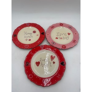 Puede incluir: Tres platos decorativos de cerámica con bordes festoneados. Cada plato presenta un diseño diferente: "Honey Bunch", "Love a bug" y "Sweet Heart". Los platos tienen bordes rojos y rosas con detalles de corazones y flores.