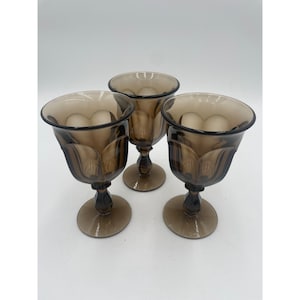 May include: Three vintage brown-tinted goblets with a tulip-shaped design. The goblets have a rounded bowl, a slender stem, and a circular base. The glass has a smoky, translucent appearance, and the goblets are arranged on a white surface.