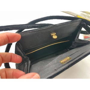 May include: Black velvet handbag with a gold clasp and a long, black cord strap. The interior is lined with black fabric and features a gold trim. The bag is open, revealing the inside.