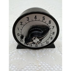 May include: A round mechanical kitchen timer with a black plastic casing and a silver-toned face. The dial features black numerals from 0 to 15, with a central black knob labeled "MARK TIME" and the printed instruction "TURN PAST 3".