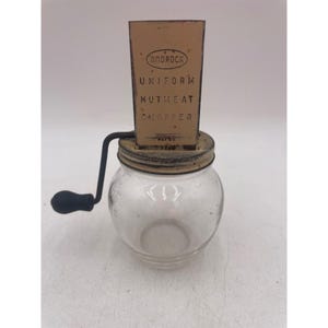 Op de afbeelding: Een antieke notenhakker met een glazen potbasis en een metalen bovenkant. De metalen bovenkant is beige met de woorden "ANDROCK UNIFORM NUTMEAT CHOPPER" erop gedrukt. Een zwarte handgreep is aan de zijkant bevestigd.