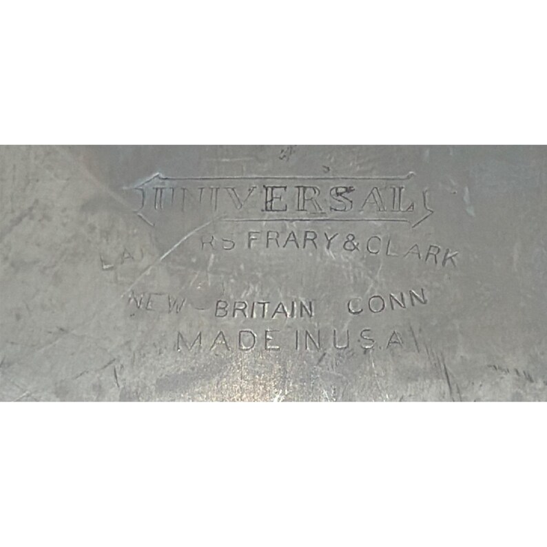 May include: A metallic surface featuring an engraved stamp that reads UNIVERSAL, LANDERS FRARY & CLARK, NEW BRITAIN CONN, and MADE IN U.S.A. The metal shows signs of wear with visible scratches and a dull, aged finish across the entire area.