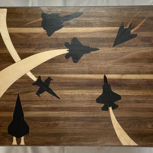 Può includere: Un tagliere in legno con finitura noce presenta cinque silhouette nere di aerei da combattimento in volo. Il tagliere ha un'intarsio in legno chiaro che forma un design curvo attorno agli aerei.