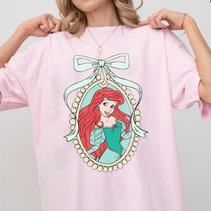 May include: Pink oversized t-shirt featuring a cartoon image of Ariel, the Little Mermaid, in a pearl-encrusted frame with a large bow.