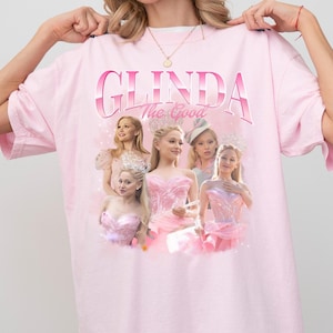 Pode incluir: Uma camiseta rosa com uma estampa gráfica de Glinda a Boa do filme Wicked. O gráfico apresenta várias imagens de Glinda em seu vestido rosa e coroa.