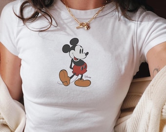 Puede incluir: Camiseta blanca recortada con una imagen de dibujos animados de Mickey Mouse con pantalones cortos rojos y zapatos negros. El texto "Disney" está impreso debajo de la imagen.