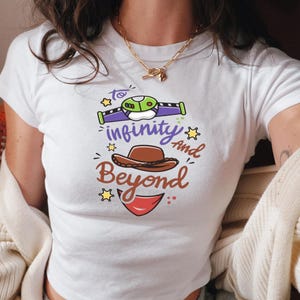 Könnte beinhalten: Weißes T-Shirt mit einem farbenfrohen Grafikdesign mit dem Satz "to infinity and beyond". Das Design umfasst ein Raumschiff, einen Cowboyhut und ein rotes Lächeln. Das Shirt ist kurzärmlig.