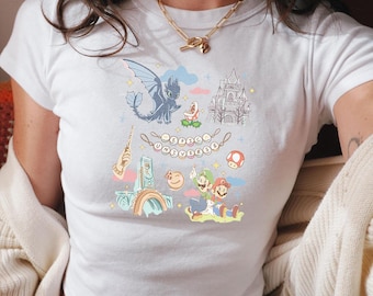 May include: White t-shirt with a colourful cartoon design featuring a dragon, castle, and video game characters. The shirt has a crew neck and short sleeves. The design includes a rainbow, mushroom, and other whimsical elements.