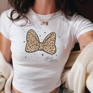 Könnte beinhalten: Weißes T-Shirt mit einem Leopardenmuster-Schleifendesign und dem Text "Minnie Mouse xoxo". Die Schleife ist beige und braun mit schwarzen Umrissen. Das T-Shirt hat einen Rundhalsausschnitt und kurze Ärmel.