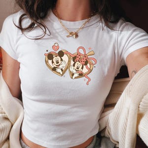 Op de afbeelding: Wit T-shirt met een retro afbeelding van Mickey en Minnie Mouse in hartvormige medaillons. Het ontwerp bevat een roze strik, sterren en een hartvormig slot. Het T-shirt heeft korte mouwen en een ronde hals.