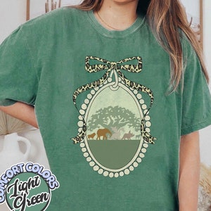 Könnte beinhalten: Ein grünes T-Shirt mit einer Vintage-Illustration einer Gruppe von Tieren in einer Waldszene. Die Illustration ist von einem Perlenrand und einer Schleife mit Leopardenmuster umrahmt.