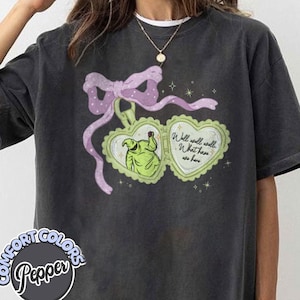 Könnte beinhalten: Dunkelgraues Comfort Colors T-Shirt mit einem herzförmigen Design, das eine grüne Figur und den Text "Well, well, well, What have we here" zeigt. Über dem Design befinden sich eine lila Schleife und ein Band.
