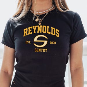 May include: Black t-shirt featuring the text "REYNOLDS SENTRY" in gold. The design includes the year "2000" and "EST." The shirt has a crew neck and is likely made of cotton.