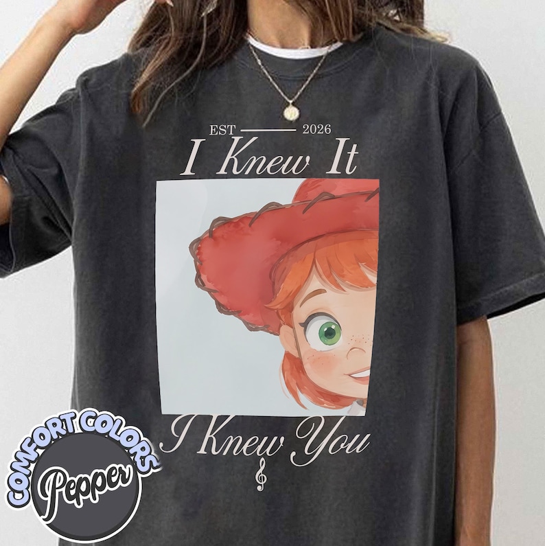 May include: A charcoal grey t-shirt featuring a graphic print of a cartoon character with red hair and a red hat. The text reads EST 2026 I Knew It I Knew You with a treble clef symbol. The shirt has a crew neckline and short sleeves.