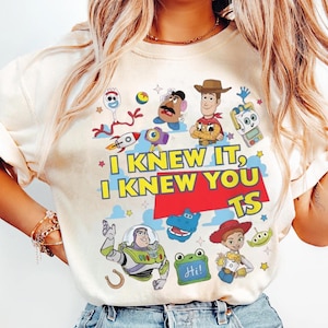 K&ouml;nnte beinhalten: Ein cremefarbenes T-Shirt mit einem bunten Grafikdruck von Toy Story-Figuren wie Woody, Buzz Lightyear, Jessie, Forky und Mr. Potato Head. Der Text lautet "I KNEW IT, I KNEW YOU TS" in fetten gelben Buchstaben auf einem roten Hintergrund.