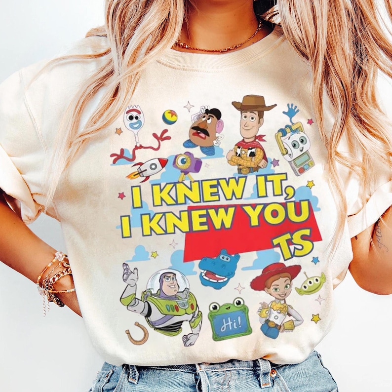K&ouml;nnte beinhalten: Ein cremefarbenes T-Shirt mit einem bunten Grafikdruck von Toy Story-Figuren wie Woody, Buzz Lightyear, Jessie, Forky und Mr. Potato Head. Der Text lautet "I KNEW IT, I KNEW YOU TS" in fetten gelben Buchstaben auf einem roten Hintergrund.