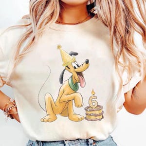 Op de afbeelding: Een crèmekleurig t-shirt met een afbeelding van een gele tekenfilmhond die een gele feesthoed en een groene halsband draagt. De hond steekt zijn tong uit en staat naast een kleine bruine verjaardagstaart met een brandende kaars in de vorm van een 6.