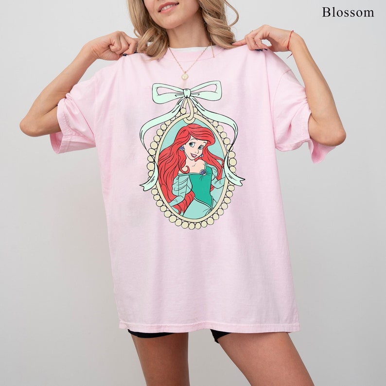 May include: Pink oversized t-shirt featuring a cartoon image of Ariel, the Little Mermaid, in a pearl-encrusted frame with a large bow.