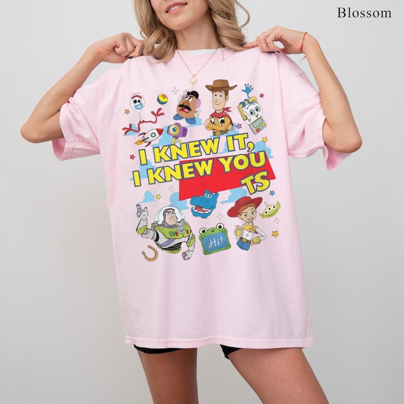 K&ouml;nnte beinhalten: Ein hellrosa T-Shirt mit einem Grafikdruck verschiedener animierter Figuren, darunter ein Weltraum-Ranger, ein Cowboy und ein Cowgirl. Der Text lautet I KNEW IT, I KNEW YOU TS. Das Design enth&auml;lt bunte Sterne, eine Rakete und kleine Spielzeuge.