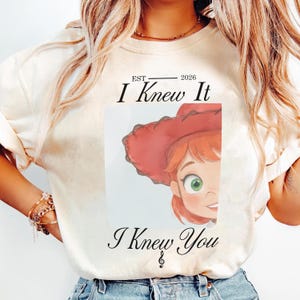 May include: A cream-coloured short-sleeve t-shirt featuring a graphic of a cartoon character with red hair and a red hat. The text reads EST 2026 I Knew It I Knew You with a treble clef symbol below the text. The design is centred on the chest.