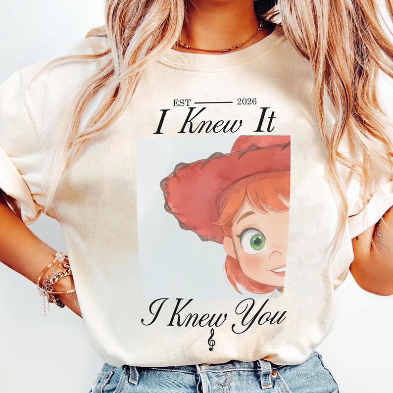 May include: A cream-coloured short-sleeve t-shirt featuring a graphic of a cartoon character with red hair and a red hat. The text reads EST 2026 I Knew It I Knew You with a treble clef symbol below the text. The design is centred on the chest.