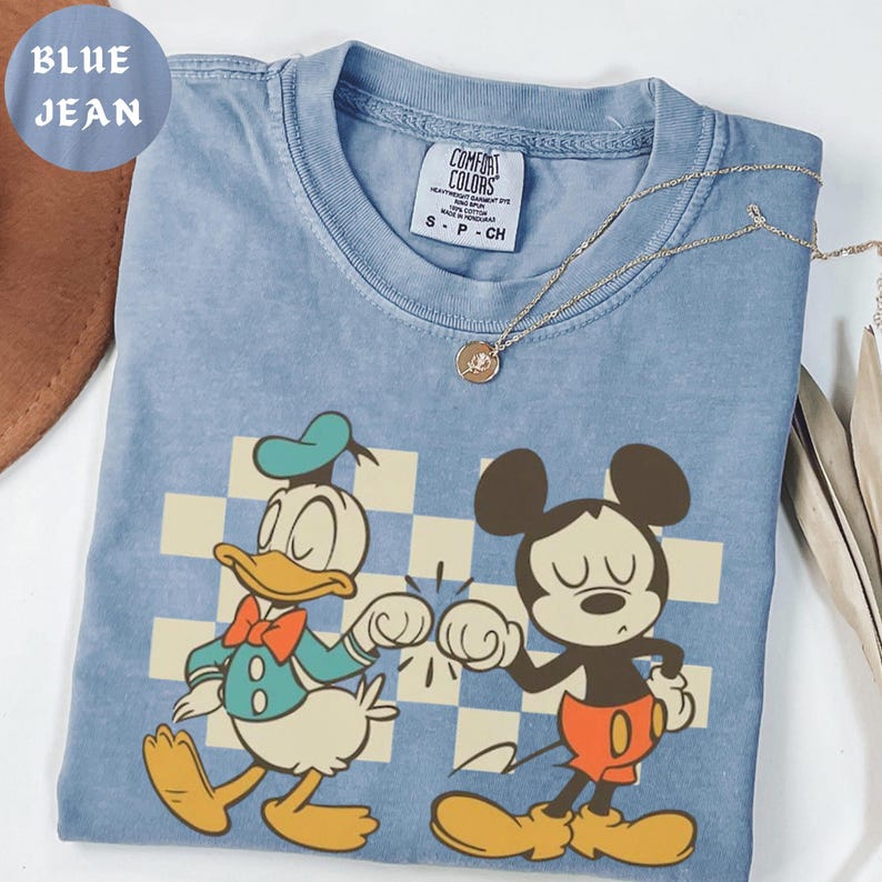 Puede incluir: Camiseta azul vaquero Comfort Colors con Donald Duck y Mickey Mouse chocando los pu&ntilde;os. La camiseta tiene un fondo a cuadros y un collar dorado. La camiseta es talla S-P-CH.