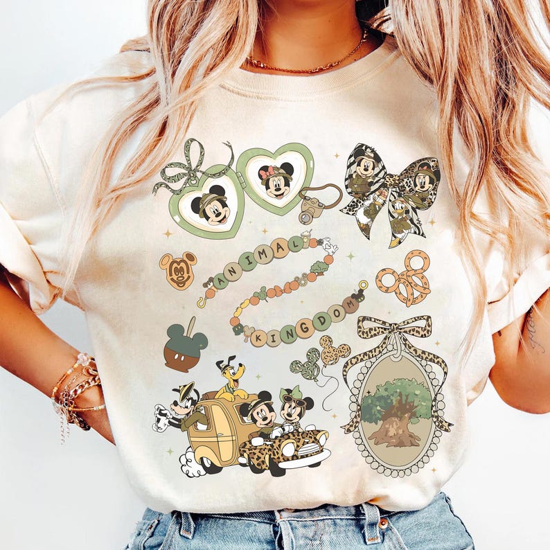 May include: Cream-colored t-shirt featuring various Disney-themed graphics. Designs include Mickey and Minnie Mouse, a beaded necklace spelling "ANIMAL KINGDOM", and a car with characters. The shirt has a crew neck and short sleeves.