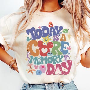 May include: A white t-shirt featuring a colourful graphic design that reads "Today is a Core Memory Day". The design incorporates various Disney characters and symbols, including Mickey Mouse, the Disney castle, and characters from the film Inside Out.