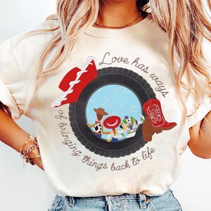 以下が含まれることがあります： 黒いタイヤのグラフィックが描かれたクリーム色のTシャツ。タイヤの中には3人の漫画キャラクターのシーンがあり、赤いカウボーイハットと赤いカウボーイブーツがタイヤの上に配置されています。テキストには「Love has ways of bringing things back to life」と書かれています。