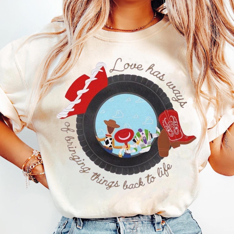 以下が含まれることがあります： 黒いタイヤのグラフィックが描かれたクリーム色のTシャツ。タイヤの中には3人の漫画キャラクターのシーンがあり、赤いカウボーイハットと赤いカウボーイブーツがタイヤの上に配置されています。テキストには「Love has ways of bringing things back to life」と書かれています。