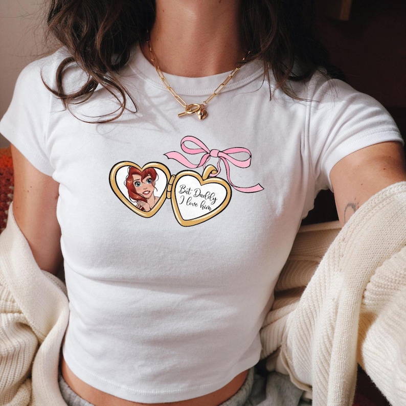 Puede incluir: Camiseta blanca con un medall&oacute;n dorado que presenta una imagen de dibujos animados de una mujer con cabello rojo. El medall&oacute;n tiene una cinta rosa y el texto "But Daddy I love him".