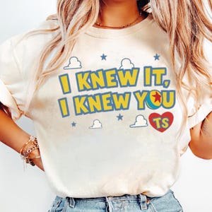 May include: A cream-coloured t-shirt featuring the text I KNEW IT, I KNEW YOU in yellow with blue outlines. The letter O in YOU is replaced by a colourful ball graphic. A red heart with the letters TS and blue star and white cloud icons surround it.