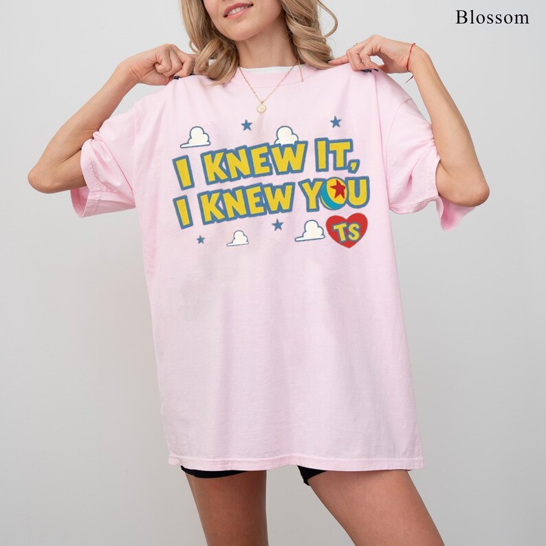 May include: A light pink t-shirt featuring the yellow text I KNEW IT, I KNEW YOU, a red heart with the letters TS, and blue star and white cloud graphics. The design is centred on the chest of the garment, which has short sleeves and a crew neck.