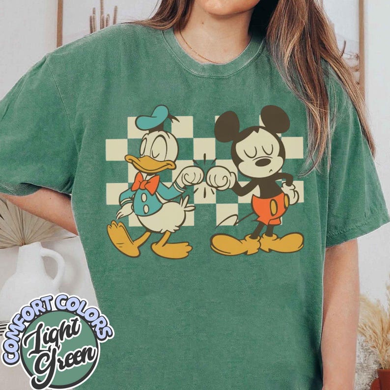 Puede incluir: Camiseta verde claro con Donald Duck y Mickey Mouse. Donald Duck lleva un sombrero azul y una pajarita, y Mickey Mouse lleva pantalones cortos rojos. La camiseta tiene un fondo a cuadros y las palabras "Comfort Colors Light Green".