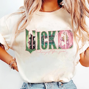 Op de afbeelding: Cr&egrave;mekleurig T-shirt met het woord "WICKED" in een groen en roze ontwerp. De letters zijn gestileerd met afbeeldingen van personages uit de musical. De zin "changed for good" staat onder de hoofdtekst in een scriptletter.