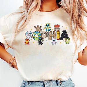 May include: Cream-colored crew neck sweatshirt with the name "SOPHIA" in colorful, dragon-themed letters. Each letter features a different dragon design, with stars scattered around the name. The sweatshirt is a casual, comfortable style.