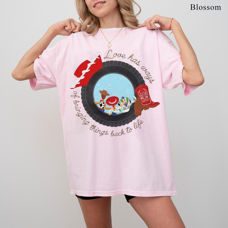 以下が含まれることがあります： 薄いピンク色のTシャツで、黒いタイヤの中に漫画のキャラクターが描かれたグラフィックが特徴です。デザインには赤いカウボーイハット、赤いカウボーイブーツ、そして円状の筆記体で「Love has ways of bringing things back to life」というテキストが含まれています。