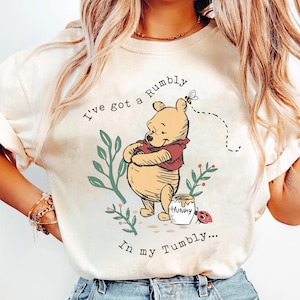 May include: Cream-colored sweatshirt featuring a cartoon illustration of Winnie the Pooh with the text "I've got a Rumbly in my Tumbly...". The design includes a honey pot, bee, and foliage.